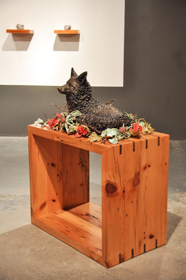 Sanctuary, cast bronze and ceramic flowers and foliage with 200-year old reclaimed local wood  A life-sized bronze fox curls up in repose encircled by a ring of ceramic flowers interwoven with thorns.  The wooden pedestal is from 200-year old wood salvaged from homes being remodelled in the artist's neighbourhood.  This work hovers between a celebration and acknowledgement of the fragility and grounding nature of home and also a memento mori.