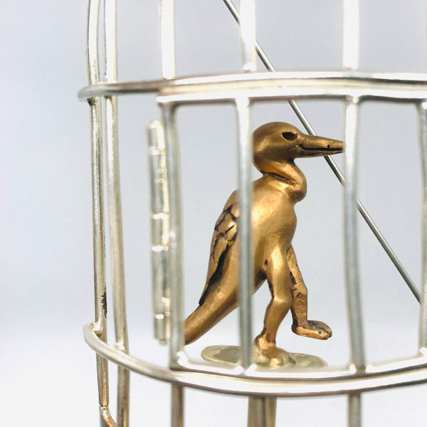 A pin of a gold duckling figurine inside a silver birdcage on a light gray background