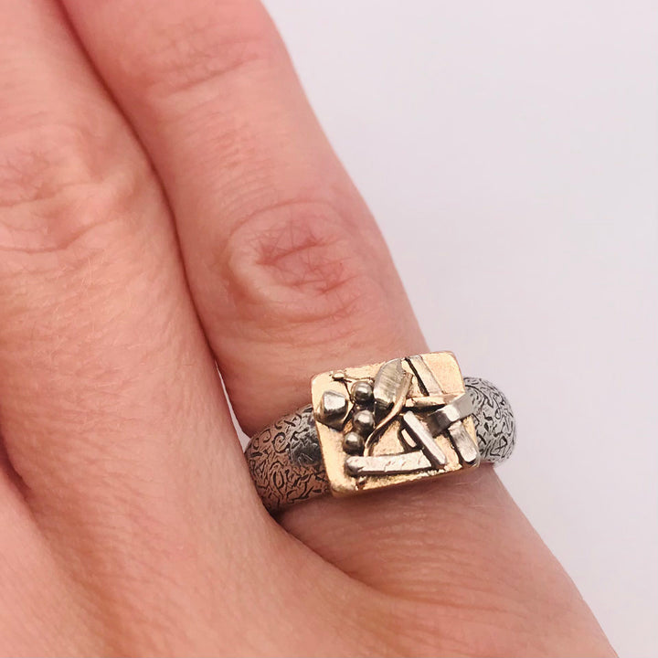 One of a kind ring in 10k yellow gold with a textured sterling silver band. Small details on the face in silver and gold suggest leaves and twigs. 2.3 x 2.1 x 0.8 cm.