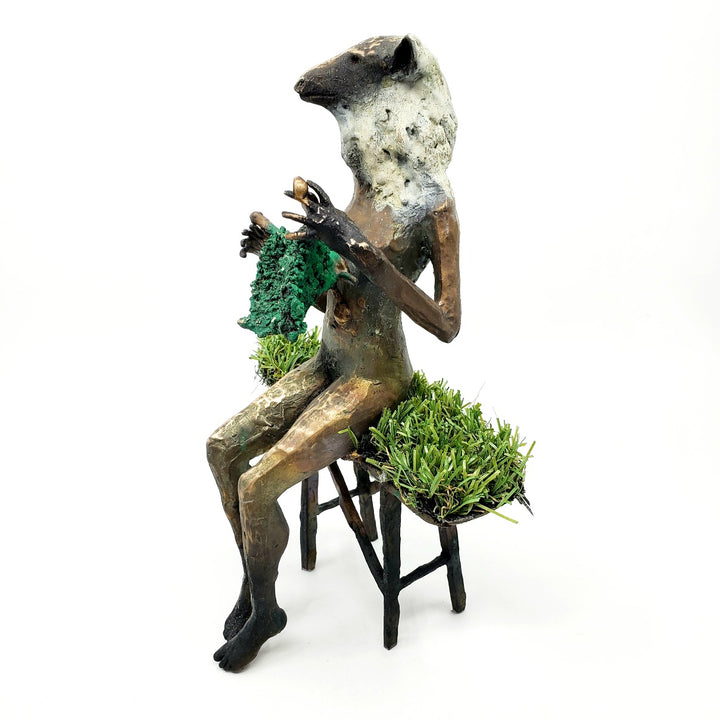 Domestic Sheep: Elsie Mae.  A bronze figure with a sheep's head and a woman's body sits on a turf-covered bench, peacefully knitting. 1 of 1.