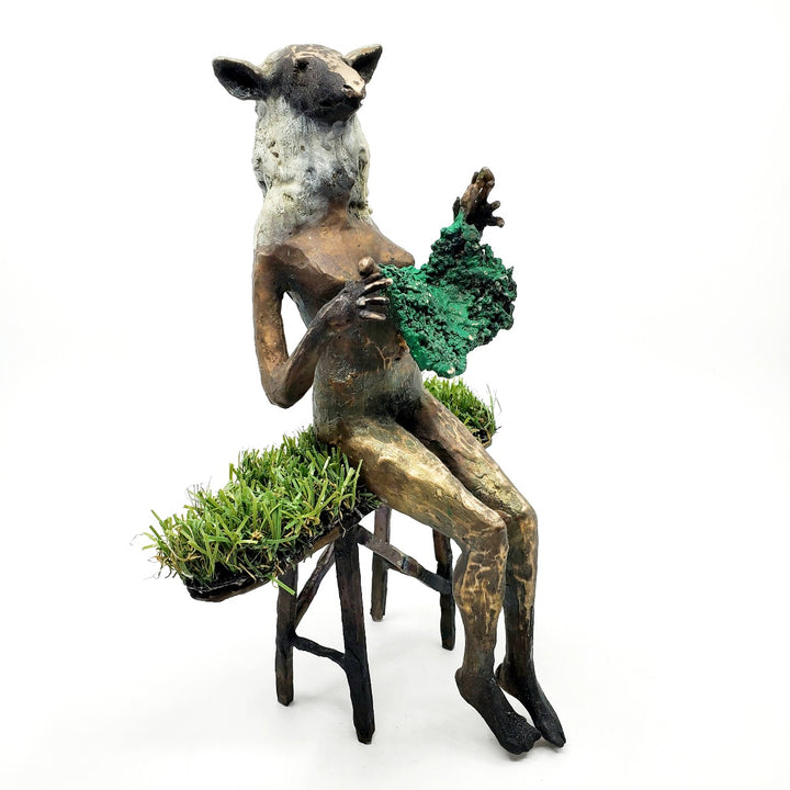 Domestic Sheep: Elsie Mae.  A bronze figure with a sheep's head and a woman's body sits on a turf-covered bench, peacefully knitting. 1 of 1.