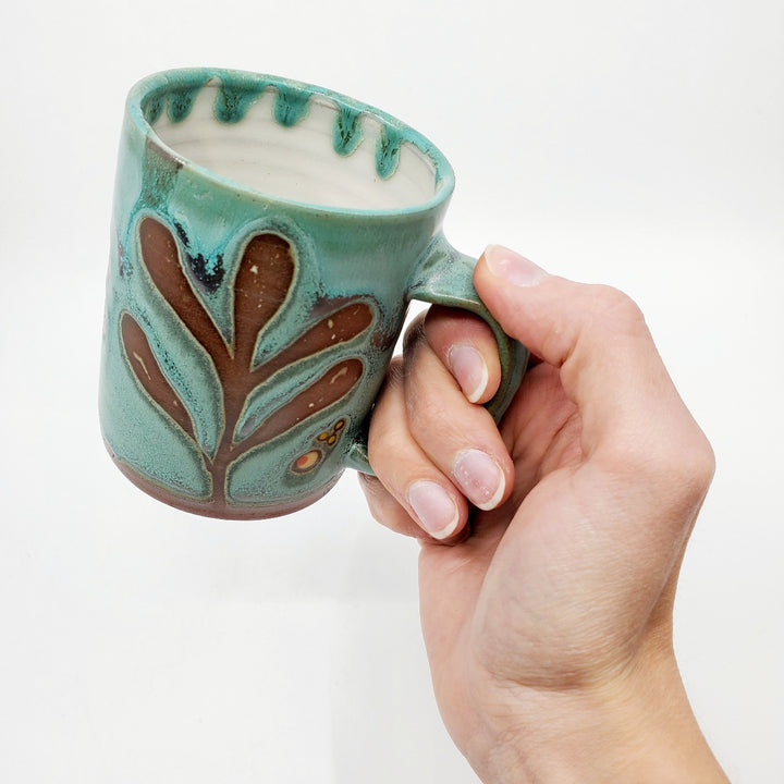 Green ceramic mug with a tactile decorated surface revealing the red clay. The interior is glazed white.  Measures approx. 8 x 10.5 x 10cm