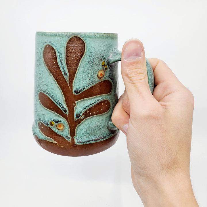 Green ceramic stein (pictured on the right) with a tactile decorated surface revealing the red clay. The interior is glazed white.  Measures approx. 8.5 x 11.5 x 12cm