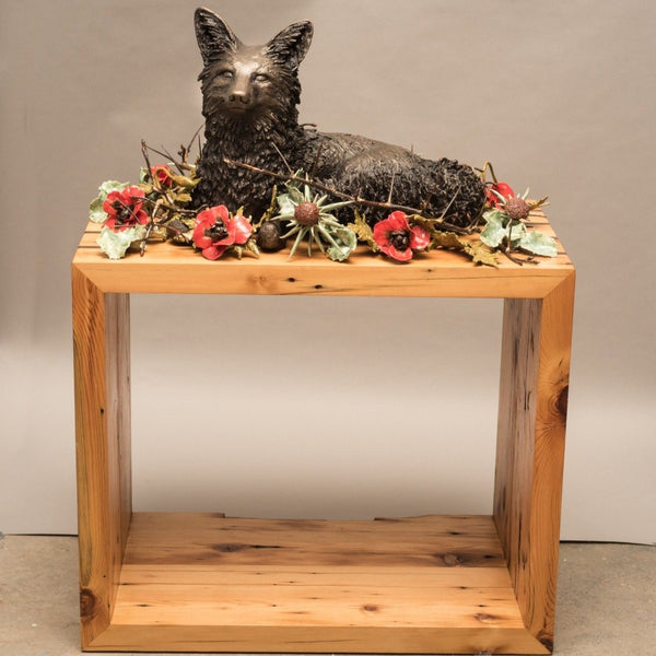 Sanctuary, cast bronze and ceramic flowers and foliage with 200-year old reclaimed local wood  A life-sized bronze fox curls up in repose encircled by a ring of ceramic flowers interwoven with thorns.  The wooden pedestal is from 200-year old wood salvaged from homes being remodelled in the artist's neighbourhood.  This work hovers between a celebration and acknowledgement of the fragility and grounding nature of home and also a memento mori.